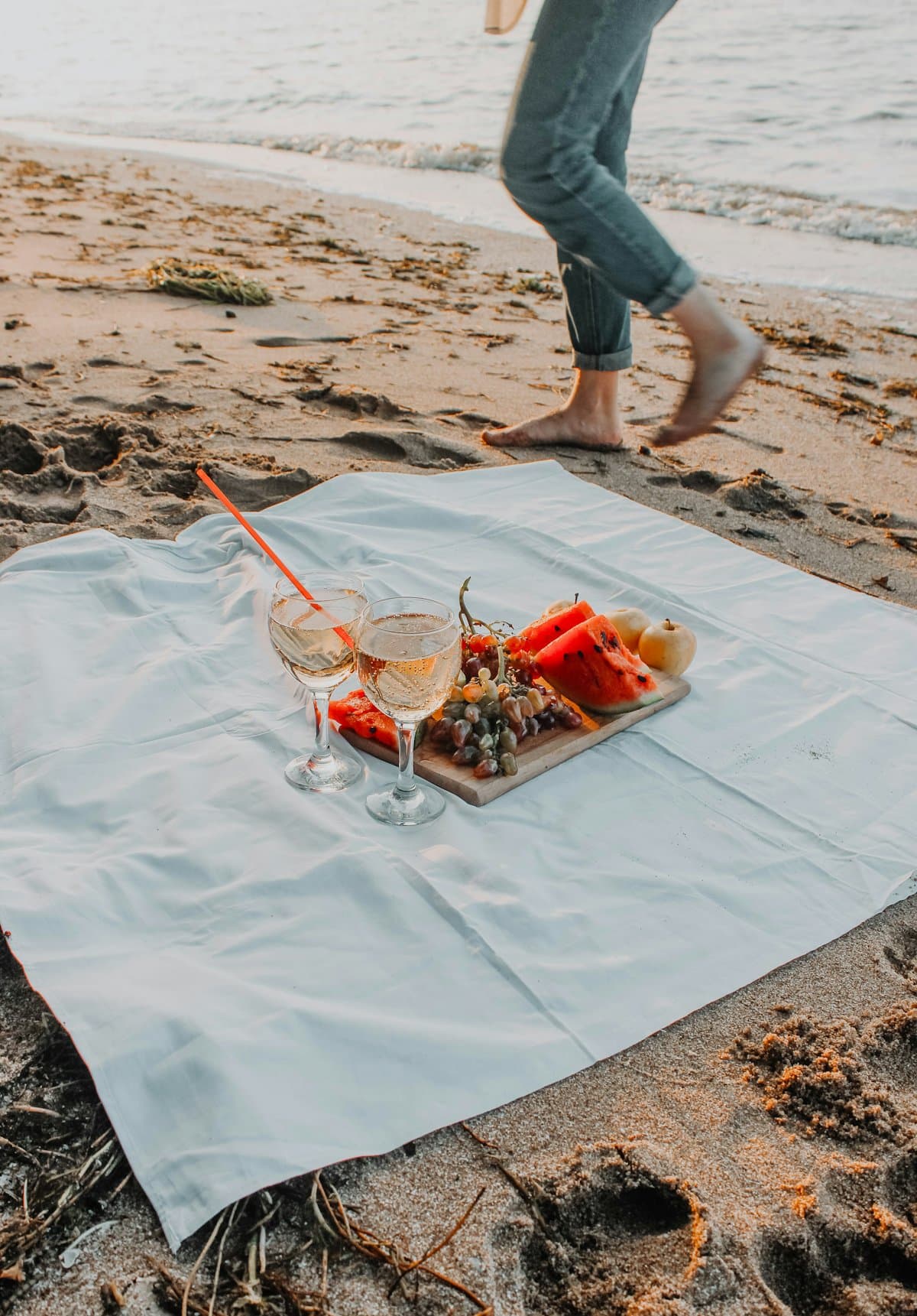 A board of fruit and two glasses of rosé on a blanket by the shore, someone walking barefoot toward the water in the golden evening light